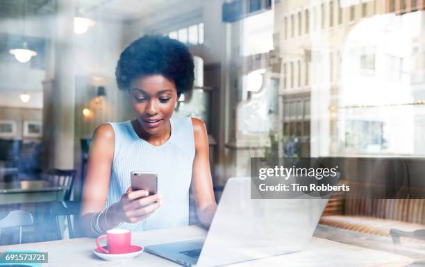 woman using smartphone with laptop in cafe - african creative with laptop working outside stockfoto's en -beelden