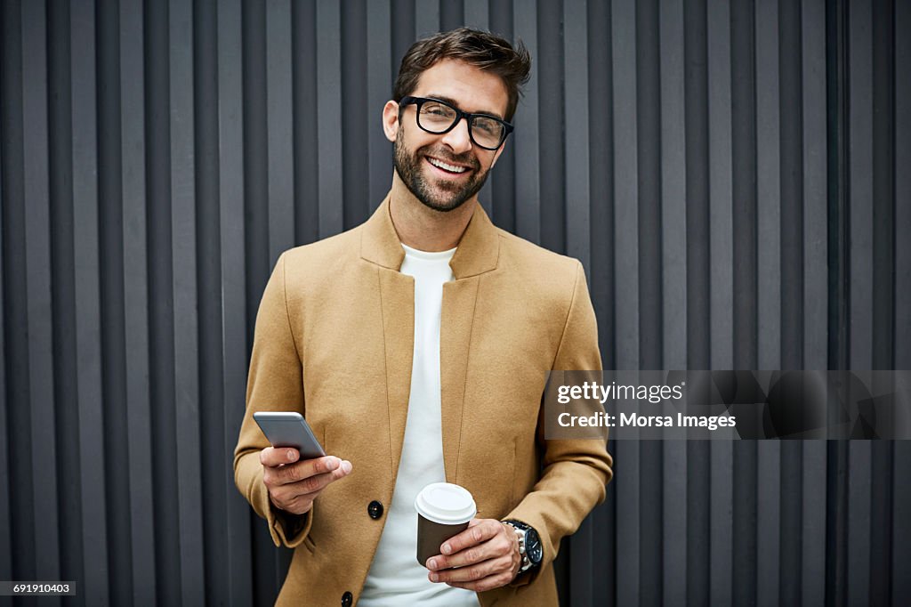 Happy businessman with smart phone and cup in city