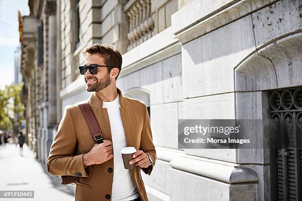 smiling businessman with cup looking away in city - gafas de sol fotografías e imágenes de stock