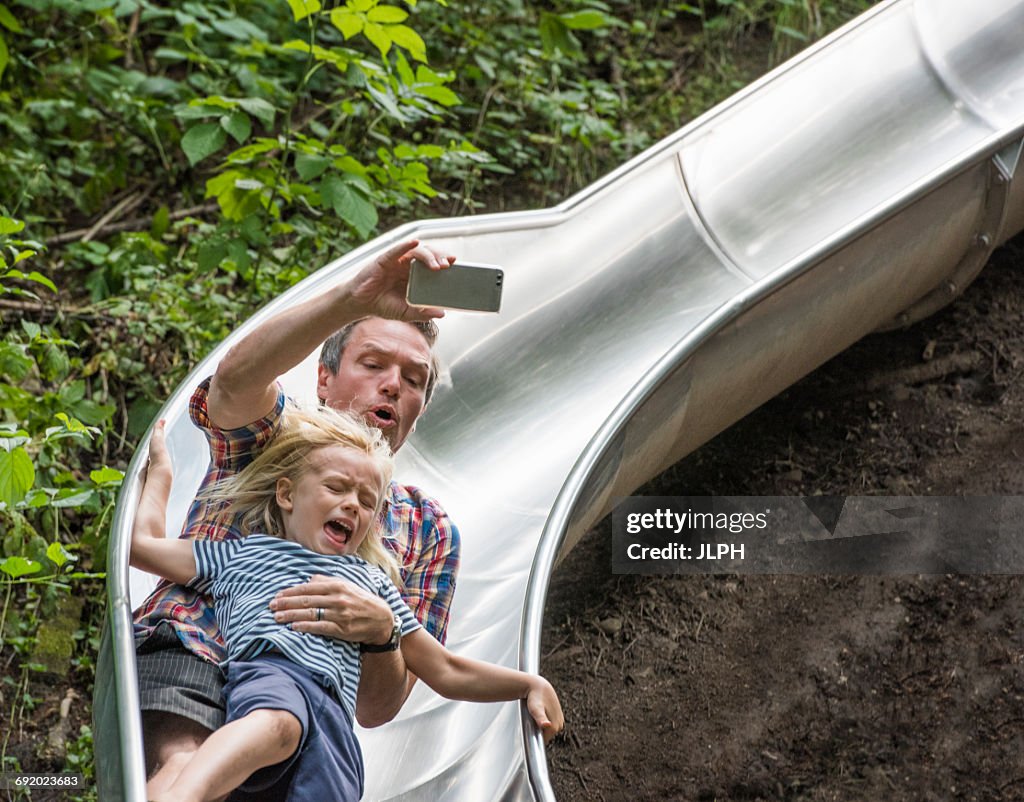 Father and son sliding down slide holding smartphone