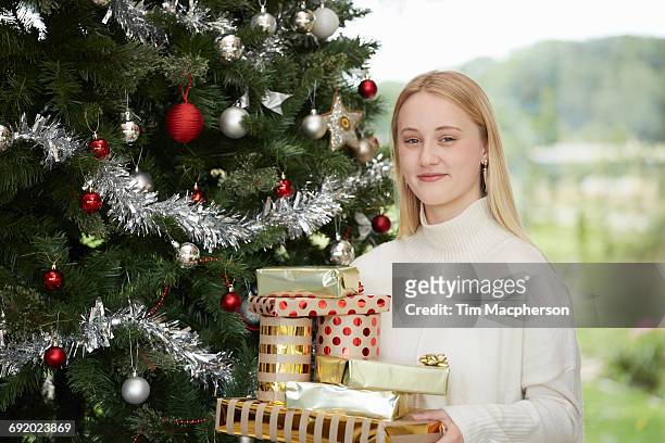 portrait of teenage girl holding stack of chrismas presents by christmas tree - halstock stock-fotos und bilder