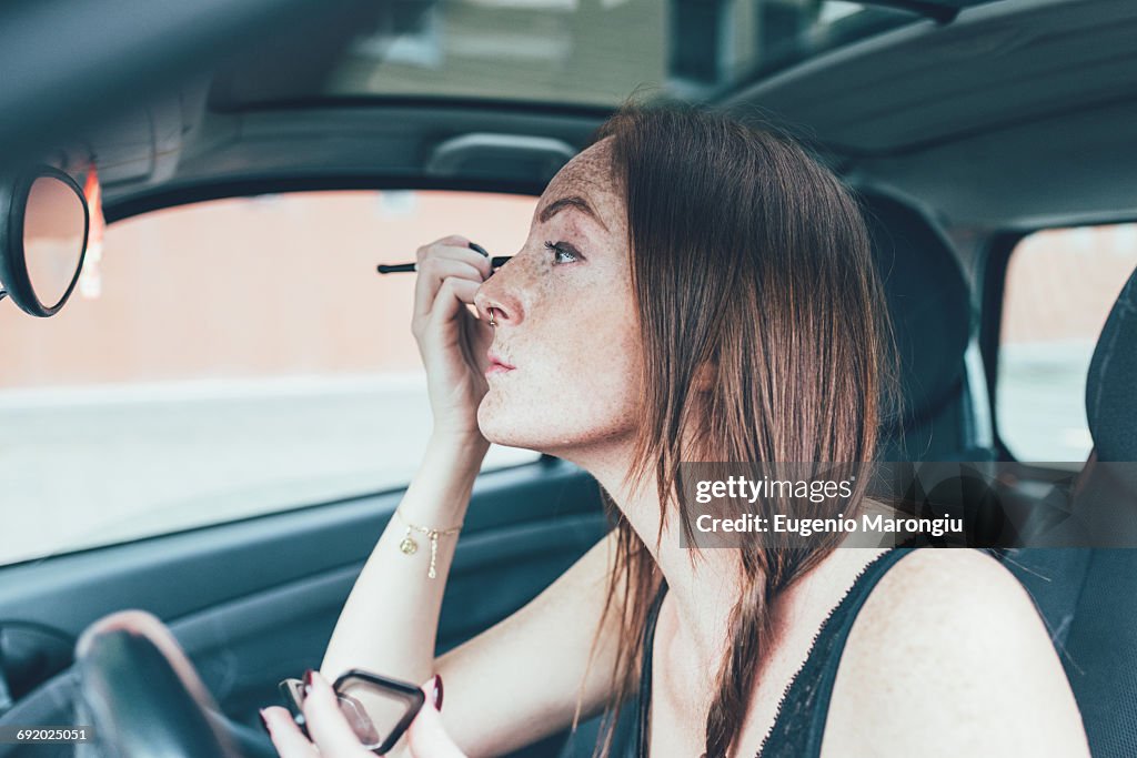 Young woman with freckles applying eyeshadow in car mirror