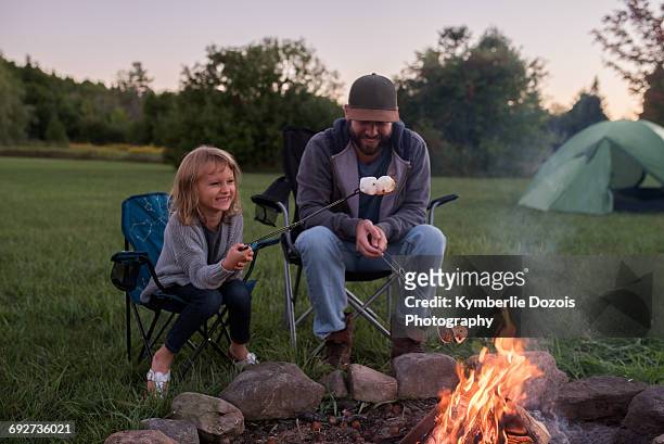father and daughter sitting beside campfire, toasting marshmallows over fire - peterborough ontario stockfoto's en -beelden