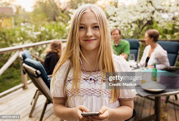 portrait of teenage girl holding smart phone at yard with family sitting in background - family back yard stockfoto's en -beelden
