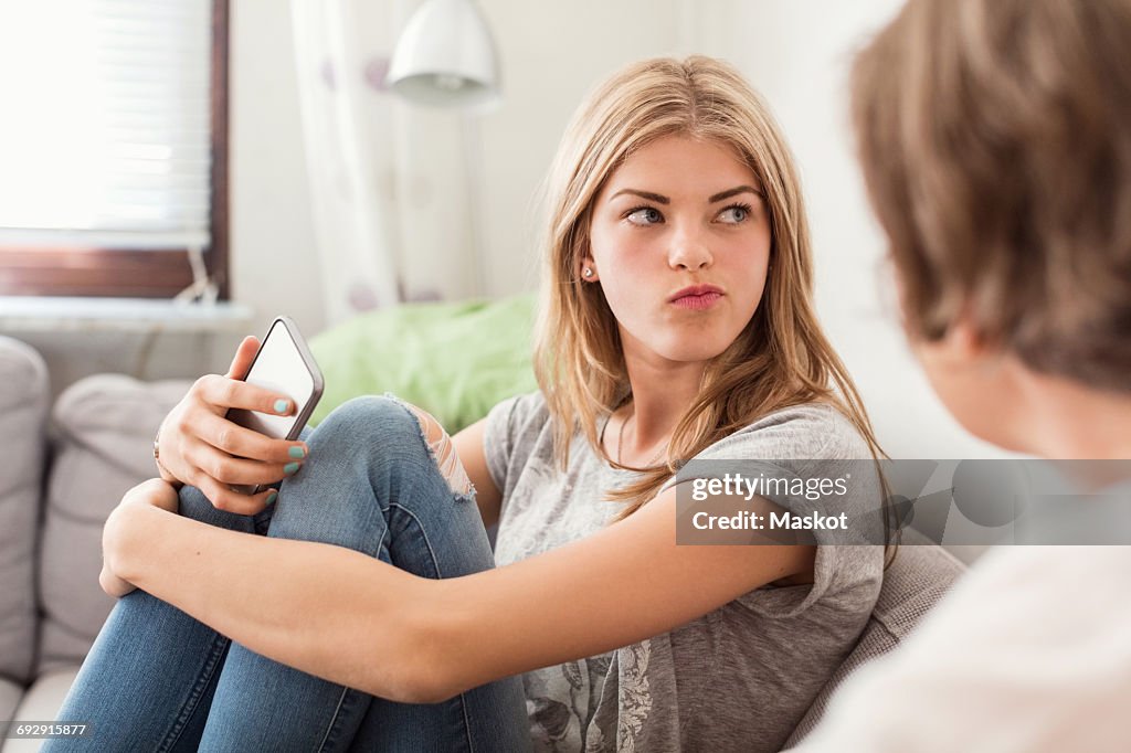 Teenage girl making faces while sitting with mother on sofa at home