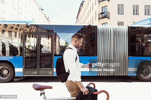 side view of businessman using smart phone while standing with bicycle against bus on street - bike headset stockfoto's en -beelden