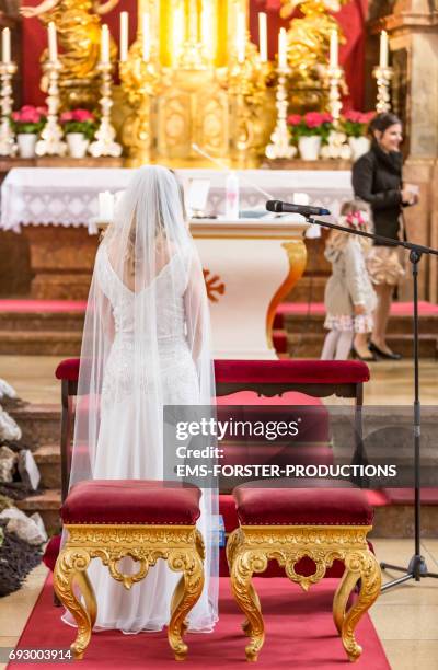 bride in wedding dress with train stands in front of the altar in a catholic church waiting for her groom and the wedding ceremony to begin - kneeler stock pictures, royalty-free photos & images