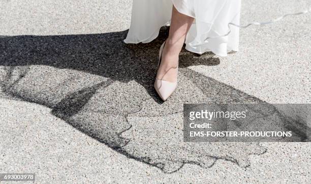 one foot of a bride wearing creme-coloured high heels pumps and her white wedding dress - shadow of the wedding dress on street top - huidkleurige schoen stockfoto's en -beelden