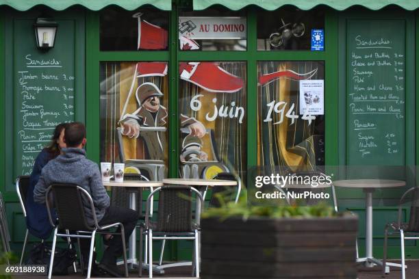 Couple having a drink in a local Cafe Bar in Sainte-Mere-Eglise with its windows decorated with D-Day themes. Tuesday 6th June is the 73rd...