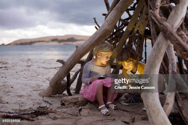 young boy and girl sit in a homemade driftwood teepee on a beautiful beach on devices/phones - mobile phone and adventure ストックフォトと画像
