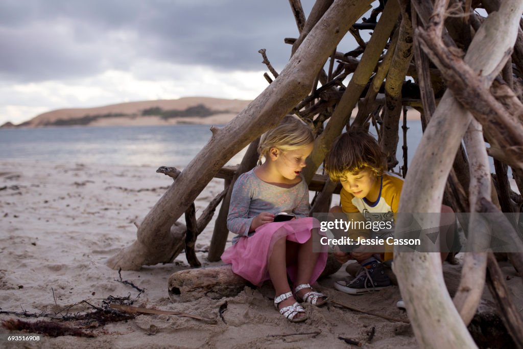 Young boy and girl sit in a homemade driftwood teepee on a beautiful beach on devices/phones