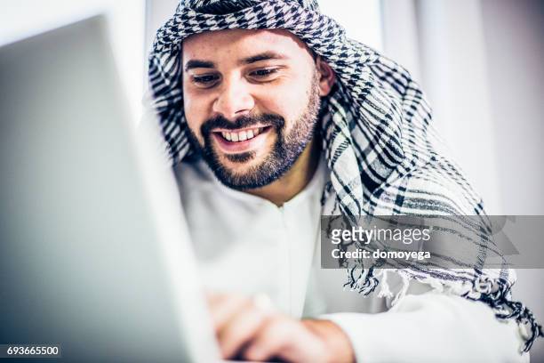 young arabian man looking at laptop in office - jalabib imagens e fotografias de stock