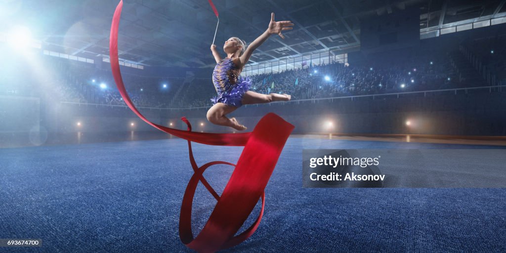 A small gymnast girl makes performance with gymnastic band on a large professional stage