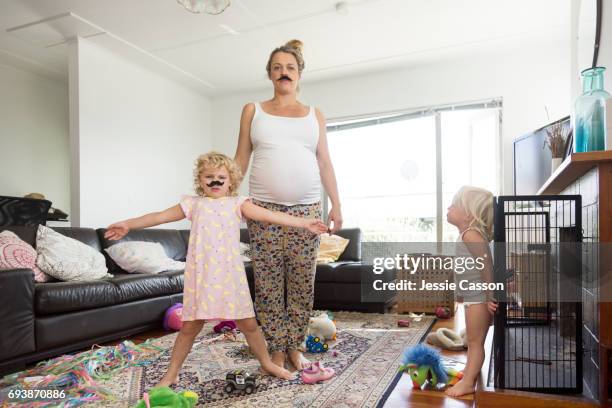 pregnant mother and two children wearing moustaches in messy living room - quirky family stockfoto's en -beelden