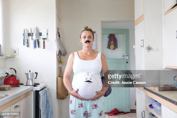 pregnant woman stands in retro kitchen wearing a moustache with a face drawn on her teeshirt also wearing a moustache - blue house red door stock pictures, royalty-free photos & images