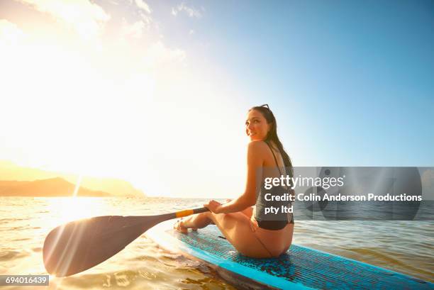 mixed race woman sitting on paddleboard in ocean - mixed race woman stock-fotos und bilder