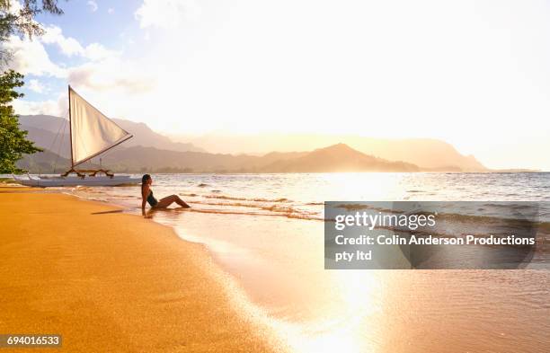 mixed race woman sitting on beach near sailboat - hawaii fun stock-fotos und bilder