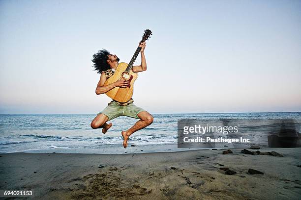 mixed race man playing guitar and jumping at beach - guitariste photos et images de collection