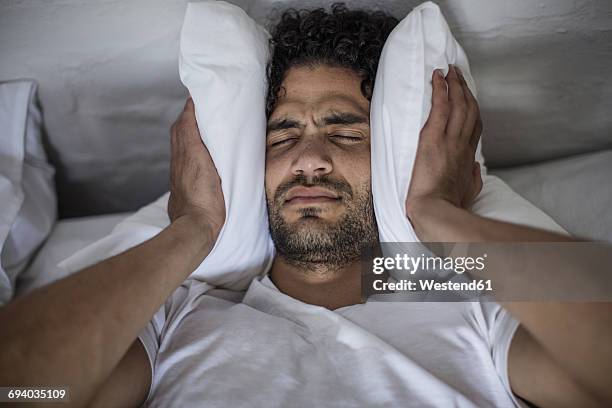 young man covering his ears with a pillow - inconveniência imagens e fotografias de stock