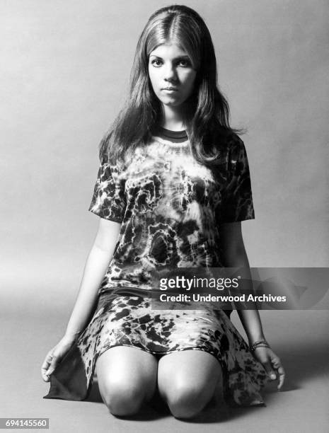 Portrait of an unidentified model as she poses, kneeling on the floor, addressed in a tie-dyed shirt with a matching scarf across her legs, 1969.
