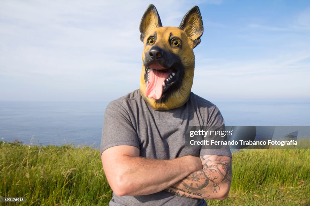 Man with tattooed arm and German Shepherd dog mask with arms crossed by the sea