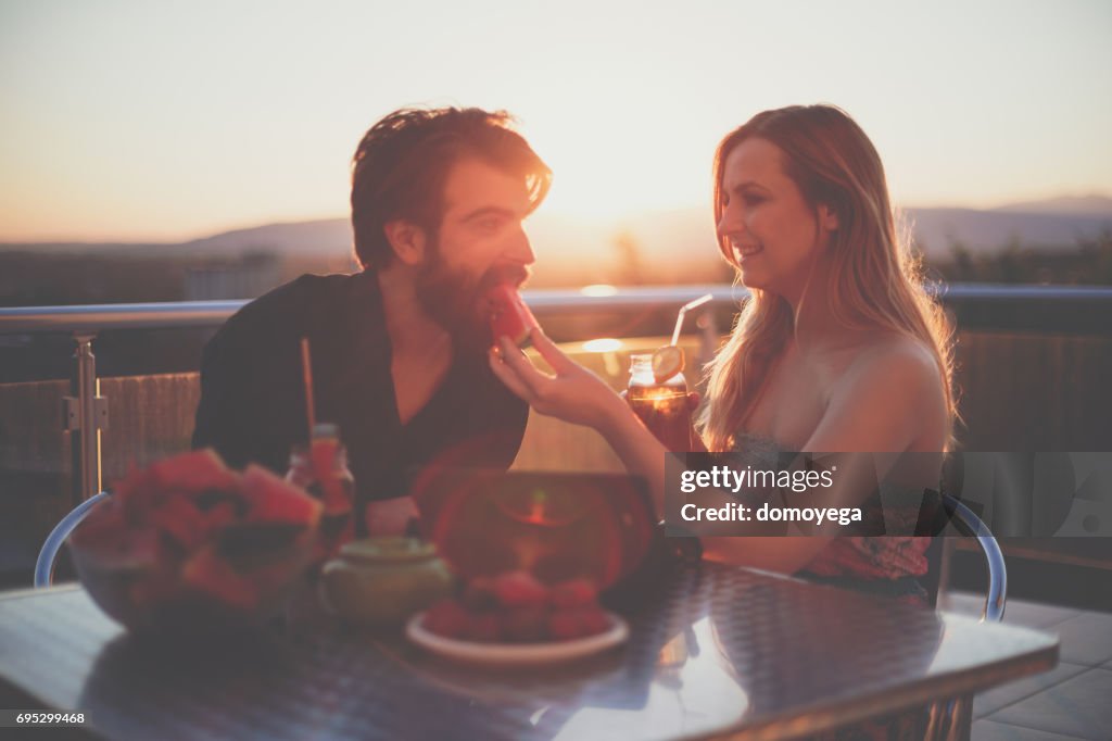 Beautiful young couple eating fruit salad and drinking smoothies on the balcony on a sunny summer day