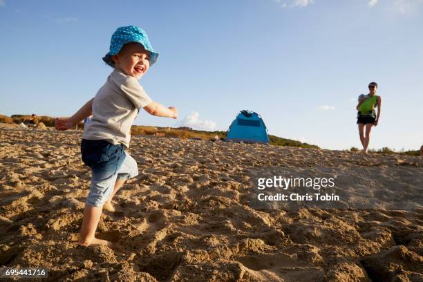 mother with baby in background watching toddler boy running on the beach - boy looking over shoulder stock pictures, royalty-free photos & images