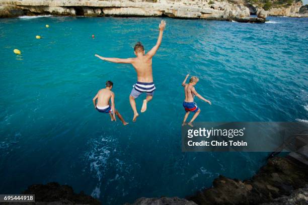three teenage boys jumping into blue water with arms outstretched - majorca 個照片及圖片檔