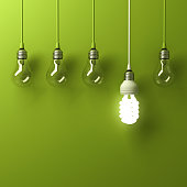 One hanging energy saving light bulb glowing different standing out from unlit incandescent bulbs with reflection on green background, leadership and different creative idea concept