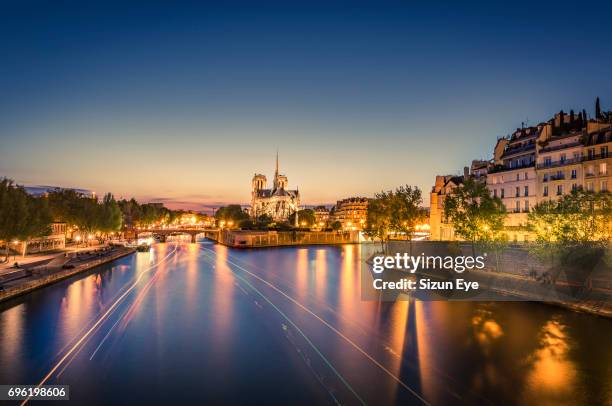 seine riverfront with notre-dame cathedral and boat light trails on the water at sunset in paris, france. - church color light paris stockfoto's en -beelden