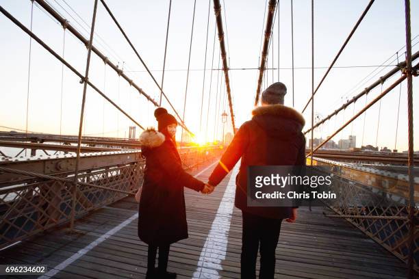 Couple in winter coats and hats holding hands on Brooklyn Bridge at sunset