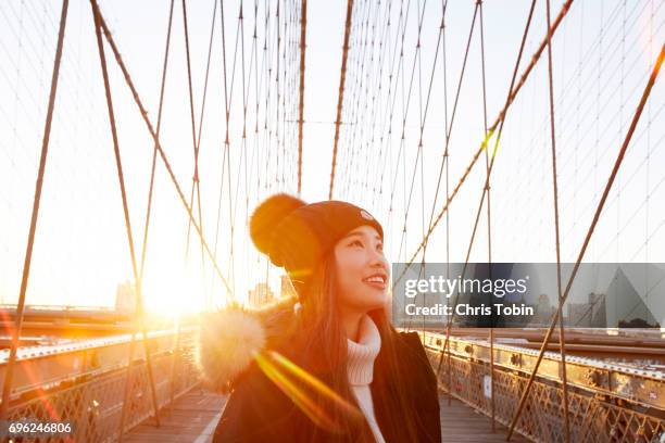 Woman in winter clothing smiling and looking up with sun in background on Brooklyn Bridge