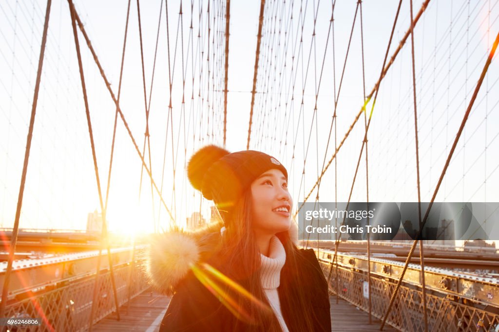 Woman in winter clothing smiling and looking up with sun in background on Brooklyn Bridge