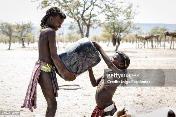 Zemba girl helping another girl drinking from a jerry can. Zembas are a bantu tribe family of the Himbas who migrated into what today is Namibia a...