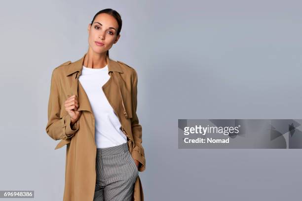 confident young woman holding brown trench coat - trench coat stock pictures, royalty-free photos & images