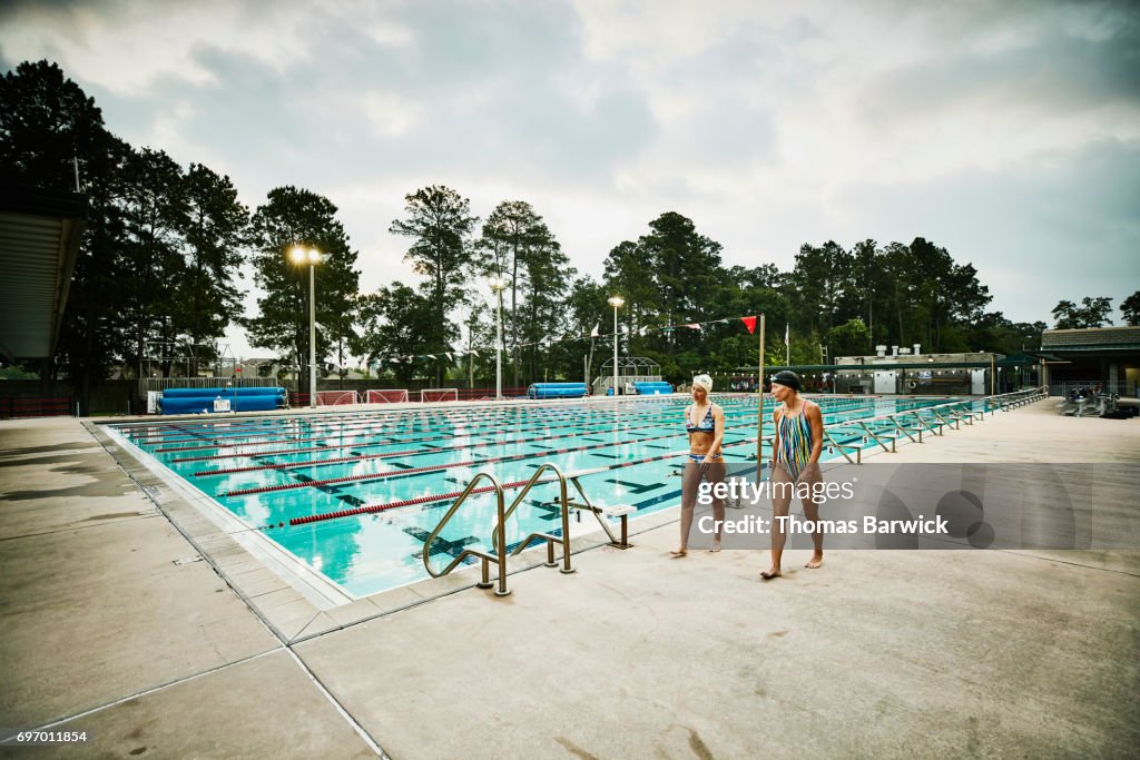 Mature female friends in discussion while walking on outdoor pool deck before early morning workout