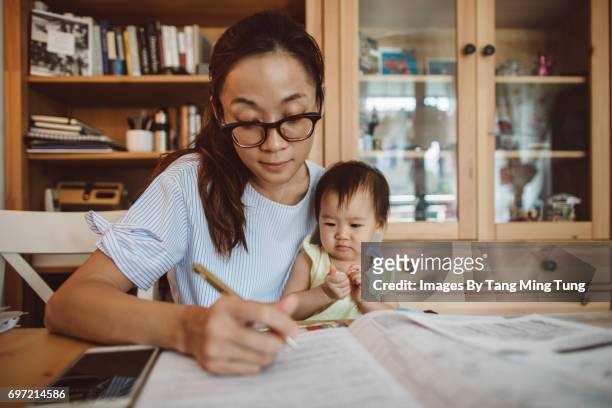 pretty young mom filling up forms and working on documents at home while holding her baby. - leanincollection working mom fotografías e imágenes de stock