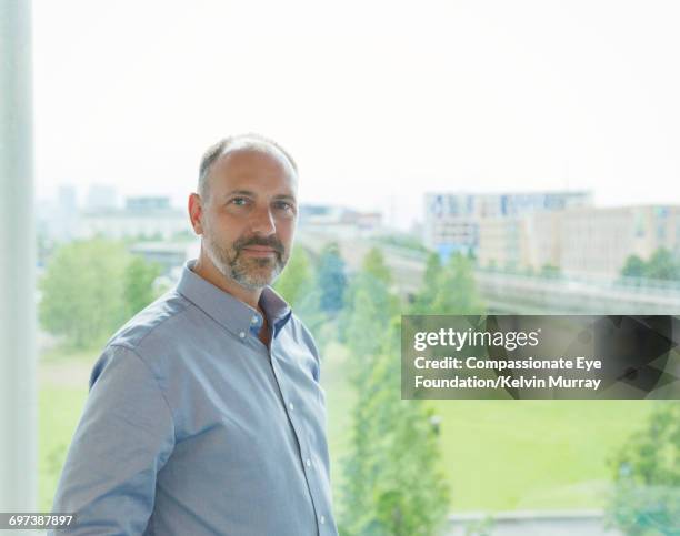 portrait of businessman standing by office window - tre quarti anteriore foto e immagini stock