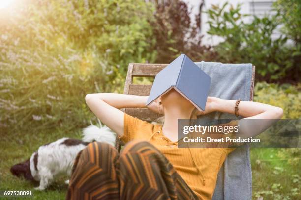 beautiful young woman covering her face with a book and relaxing in the domestic garden, munich, bavaria, germany - zonnestoel stockfoto's en -beelden
