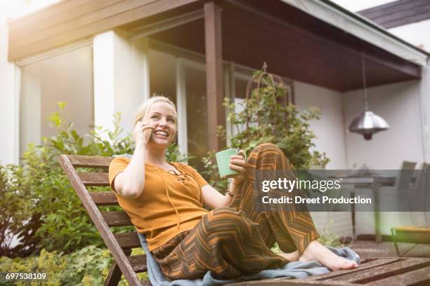 beautiful young woman having cup of tea and talking on smartphone in the domestic garden, munich, bavaria, germany - bavarian man in front of house stock-fotos und bilder