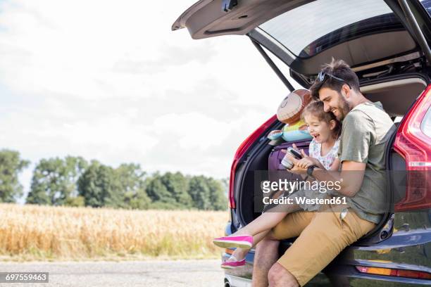 father and daughter sitting in open car boot looking at camera at break of a road trip - car family stock-fotos und bilder