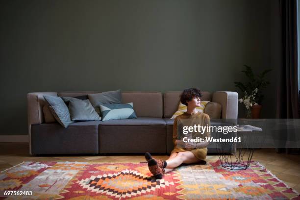 young woman sitting on the floor in her living room looking through window - sitting on the floor stock-fotos und bilder