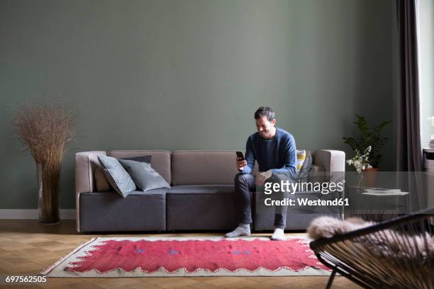 man sitting on couch in his the living room looking at smartphone - divano foto e immagini stock