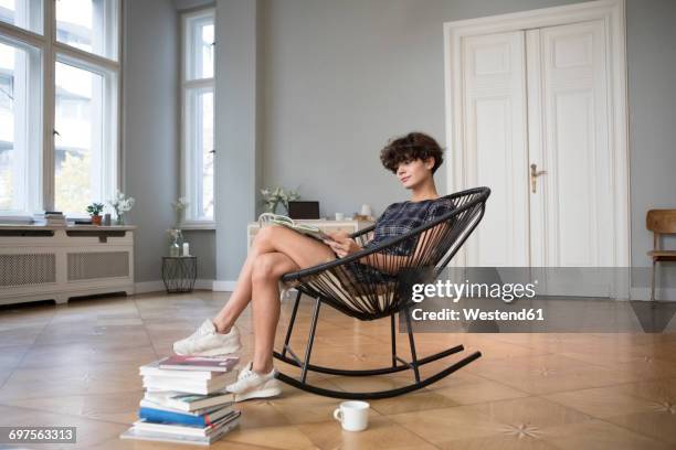 young woman sitting on rocking chair at home reading a book - cadeira de balanço - fotografias e filmes do acervo