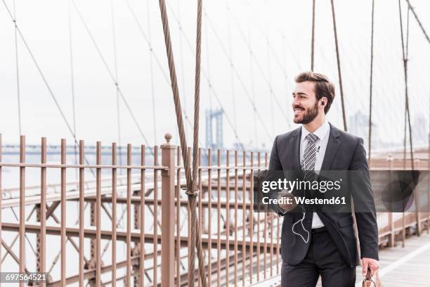 usa, new york city, smiling businessman with cell phone and earbuds on brooklyn bridge - walking on brooklyn bridge stock pictures, royalty-free photos & images