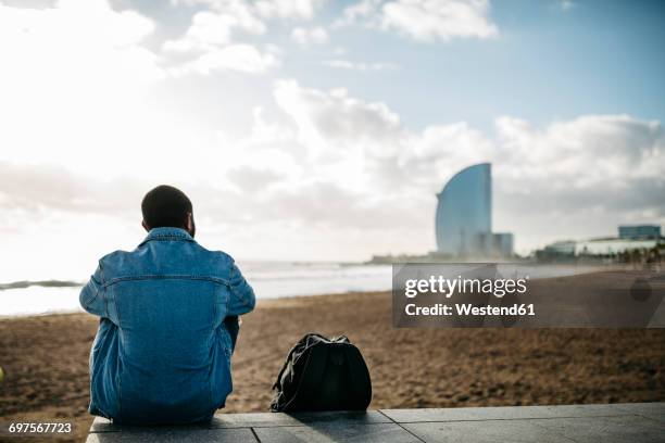 spain, barcelona, back view of young traveler sitting at the beach - man in denim jacket stock pictures, royalty-free photos & images