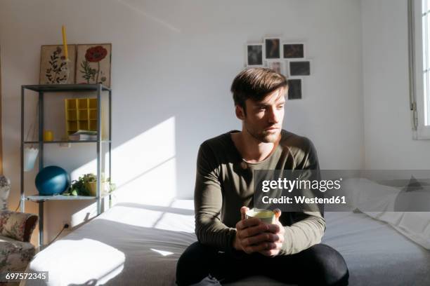 unhappy young man sitting on bed with cup of coffee - lonely man stockfoto's en -beelden