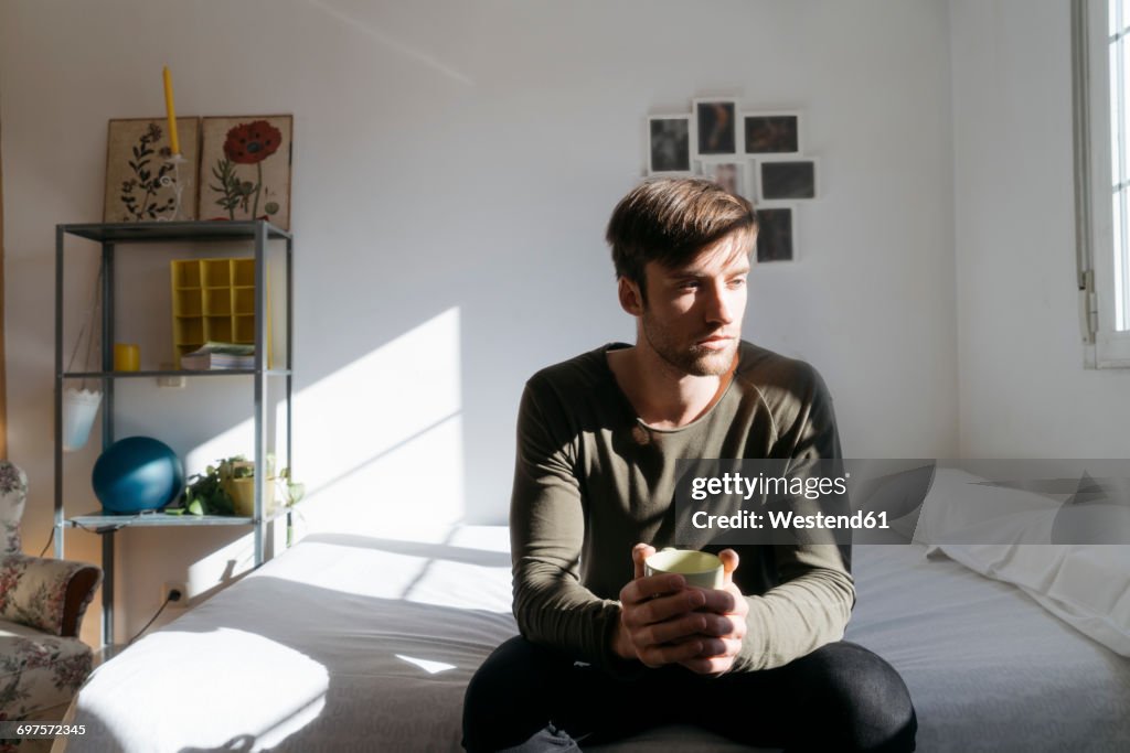 Unhappy young man sitting on bed with cup of coffee