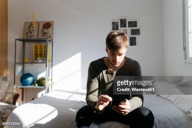 young man sitting on bed looking at his smartphone - sadness man stock pictures, royalty-free photos & images