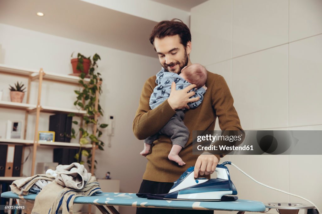 Father ironing and holding his baby at home